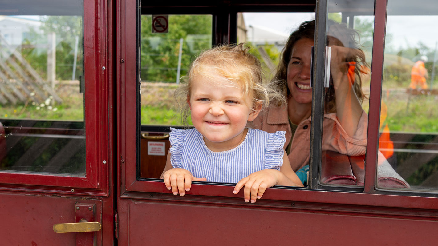 Little girl poking her head out of a train carriage, mum in background